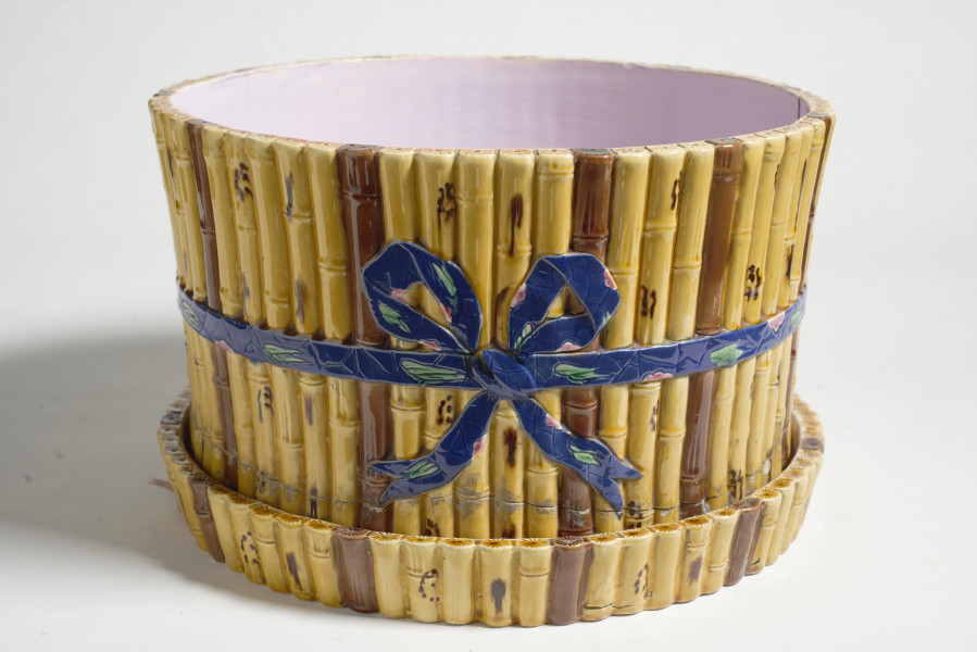 cache-pôt to duże naczynie cylindryczne, o grubych ściankach, z podstawką. Zewnętrzna powierzchnia ścianek modelowana jest na kształt wiązek łodyg bambusowych przewiązanych wstążką z kokardą. Łodygi malowane są naturalistycznie, w kolorze żółtym i brązowym. Donica i podstawka pokryte są wewnątrz polewą w kolorze liliowym. 