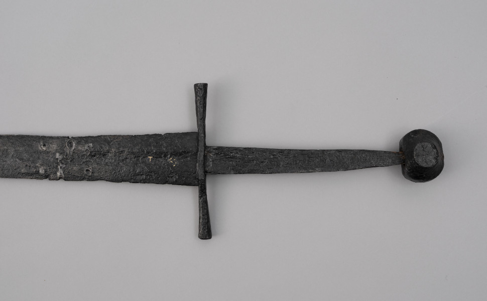 miecz typu XIVa J2 (Oakeschott) - ujęcie 3/4; Miecz długi półtoraręczny. Głownia długości 95 cm i szer. u nasady 5,6 cm, prosta dwusieczna zbieżna ku sztychowi, łączona wtórnie w dwóch miejscach. Pierwotnie po obu stronach głowni znajdował się napis ryty, wypełniony żółtym metalem w postaci krzyżyka oraz trzech liter, z których pierwsza jest nieczytelna, ale przypomina L lub T; druga, środkowa to D; trzecia litera to połączenie I i H.  Jelec krzyżowy o długości 16,7 cm, z rozszerzającymi się ramionami. Głowica kolista, fazowana, dwubocznie spłaszczona o wys. 4,3 cm i szer. 5,6 cm. Powierzchnia z wżerami pokorozyjnymi. Typ wg Oakeschotta: XIVa J2 (ewentualnie XIIIa).