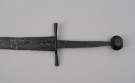 miecz typu XIVa J2 (Oakeschott) - ujęcie 3/4; Miecz długi półtoraręczny. Głownia długości 95 cm i szer. u nasady 5,6 cm, prosta dwusieczna zbieżna ku sztychowi, łączona wtórnie w dwóch miejscach. Pierwotnie po obu stronach głowni znajdował się napis ryty, wypełniony żółtym metalem w postaci krzyżyka oraz trzech liter, z których pierwsza jest nieczytelna, ale przypomina L lub T; druga, środkowa to D; trzecia litera to połączenie I i H.  Jelec krzyżowy o długości 16,7 cm, z rozszerzającymi się ramionami. Głowica kolista, fazowana, dwubocznie spłaszczona o wys. 4,3 cm i szer. 5,6 cm. Powierzchnia z wżerami pokorozyjnymi. Typ wg Oakeschotta: XIVa J2 (ewentualnie XIIIa).