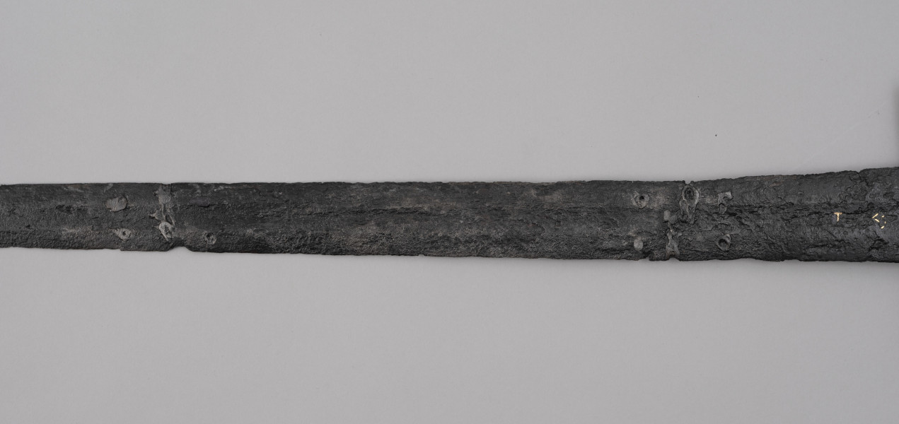 miecz typu XIVa J2 (Oakeschott) - ujęcie 3/4; Miecz długi półtoraręczny. Zbrocze długie, sięga 3/4 długości. Pierwotnie po obu stronach głowni znajdował się napis ryty, wypełniony żółtym metalem w postaci krzyżyka oraz trzech liter, z których pierwsza jest nieczytelna, ale przypomina L lub T; druga, środkowa to D; trzecia litera to połączenie I i H. Trzpień rękojeści zwężający się ku końcowi. Powierzchnia z wżerami pokorozyjnymi. Typ wg Oakeschotta: XIVa J2 (ewentualnie XIIIa).