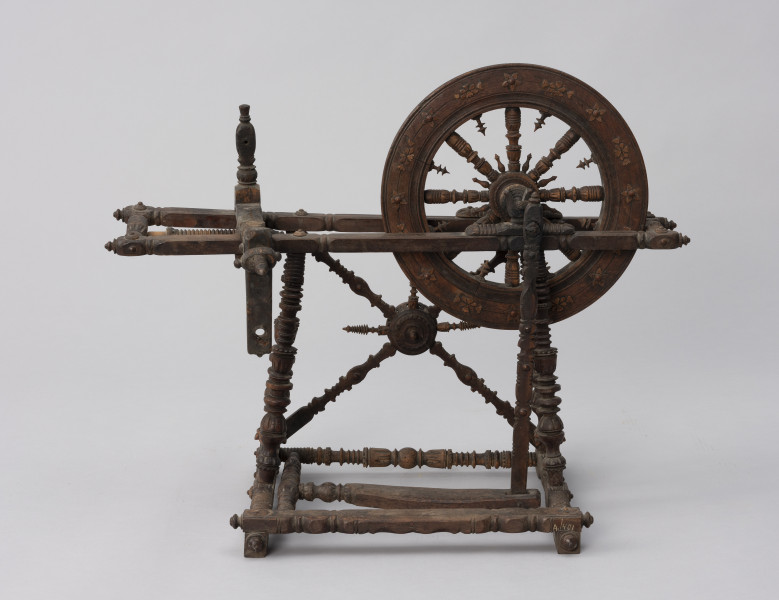 Kołowrotek poziomy - ujęcie z przodu; Kołowrotek poziomy o napędzie nożnym. Wykonany z drewna. Elementy kołowrotka łączone na wpusty. Koło napędowe o średnicy 31 cm. osadzone w podłużnej, poziomej ramie. Wykonana z metalu oś koła oparta na  wspornikach o kształcie przypominającym literę „U”. Pierwotnie na drugim końcu ramy mocowany zespół wrzeciona (zachowała się jedynie jego ruchoma podpora). Rama ustawiona na czterech, lekko pochylonych ku środkowi, toczonych na tokarce podpórkach. Pomiędzy dwiema z nich zamontowane skośnie dwa słupki, na przecięciu których znajduje się element zdobniczy - kwiat (róża?). Podpórki wsparte na prostokątnej podstawie z czterema niewielkimi nóżkami (zachowała się jedna). Pedał w postaci prostej, profilowanej na końcu deseczki połączony z korbą koła napędowego za pomocą drewnianego korbowodu. Całość bogato zdobiona.