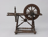 Kołowrotek poziomy - ujęcie z przodu; Kołowrotek poziomy o napędzie nożnym. Wykonany z drewna. Elementy kołowrotka łączone na wpusty. Koło napędowe o średnicy 31 cm. osadzone w podłużnej, poziomej ramie. Wykonana z metalu oś koła oparta na  wspornikach o kształcie przypominającym literę „U”. Pierwotnie na drugim końcu ramy mocowany zespół wrzeciona (zachowała się jedynie jego ruchoma podpora). Rama ustawiona na czterech, lekko pochylonych ku środkowi, toczonych na tokarce podpórkach. Pomiędzy dwiema z nich zamontowane skośnie dwa słupki, na przecięciu których znajduje się element zdobniczy - kwiat (róża?). Podpórki wsparte na prostokątnej podstawie z czterema niewielkimi nóżkami (zachowała się jedna). Pedał w postaci prostej, profilowanej na końcu deseczki połączony z korbą koła napędowego za pomocą drewnianego korbowodu. Całość bogato zdobiona.