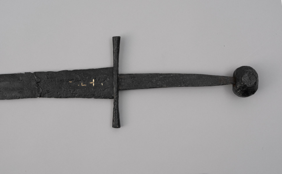 miecz typu XIVa J2 (Oakeschott) - ujęcie 3/4; Miecz długi półtoraręczny. Głownia długości 95 cm i szer. u nasady 5,6 cm, prosta dwusieczna zbieżna ku sztychowi, łączona wtórnie w dwóch miejscach. Zbrocze długie, sięga 3/4 długości. Pierwotnie po obu stronach głowni znajdował się napis ryty, wypełniony żółtym metalem w postaci krzyżyka oraz trzech liter, z których pierwsza jest nieczytelna, ale przypomina L lub T; druga, środkowa to D; trzecia litera to połączenie I i H. Jelec krzyżowy o długości 16,7 cm, z rozszerzającymi się ramionami. Głowica kolista, fazowana, dwubocznie spłaszczona o wys. 4,3 cm i szer. 5,6 cm. Powierzchnia z wżerami pokorozyjnymi. Typ wg Oakeschotta: XIVa J2 (ewentualnie XIIIa).