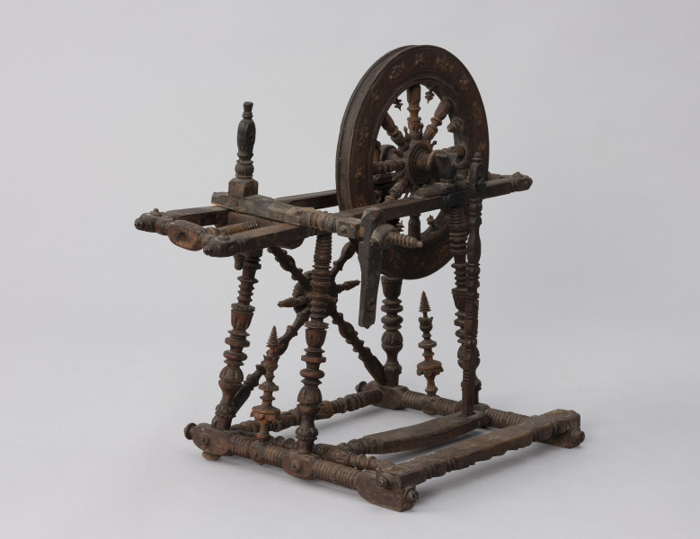 Kołowrotek poziomy - ujęcie ze skosu od tyłu; Kołowrotek  poziomy o napędzie nożnym. Wykonany z drewna. Elementy kołowrotka łączone na wpusty. Koło napędowe o średnicy 31 cm. osadzone w podłużnej, poziomej ramie. Wykonana z metalu oś koła oparta na  wspornikach o kształcie przypominającym literę „U”. Pierwotnie na drugim końcu ramy mocowany zespół wrzeciona (zachowała się jedynie jego ruchoma podpora). Rama ustawiona na czterech, lekko pochylonych ku środkowi, toczonych na tokarce podpórkach. Pomiędzy dwiema z nich zamontowane skośnie dwa słupki. Podpórki wsparte na prostokątnej podstawie z czterema niewielkimi nóżkami (zachowała się jedna). Pedał w postaci prostej, profilowanej na końcu deseczki połączony z korbą koła napędowego za pomocą drewnianego korbowodu. Całość bogato zdobiona.