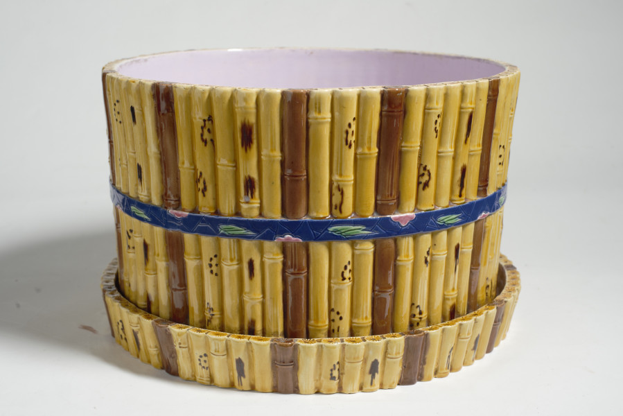 duże naczynie cylindryczne, o grubych ściankach, z podstawką. Zewnętrzna powierzchnia ścianek modelowana jest na kształt wiązek łodyg bambusowych przewiązanych wstążką z kokardą. Łodygi malowane są naturalistycznie, w kolorze żółtym i brązowym. Donica i podstawka pokryte są wewnątrz polewą w kolorze liliowym. 