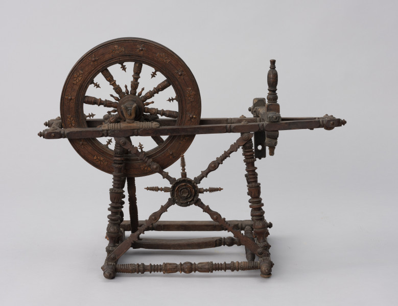 Kołowrotek poziomy - ujęcie z boku; Kołowrotek  poziomy o napędzie nożnym. Wykonany z drewna. Elementy kołowrotka łączone na wpusty. Koło napędowe o średnicy 31 cm. osadzone w podłużnej, poziomej ramie. Wykonana z metalu oś koła oparta na  wspornikach o kształcie przypominającym literę „U”. Pierwotnie na drugim końcu ramy mocowany zespół wrzeciona (zachowała się jedynie jego ruchoma podpora). Rama ustawiona na czterech, lekko pochylonych ku środkowi, toczonych na tokarce podpórkach. Pomiędzy dwiema z nich zamontowane skośnie dwa słupki, na przecięciu których znajduje się element zdobniczy - kwiat (róża?). Podpórki wsparte na prostokątnej podstawie z czterema niewielkimi nóżkami (zachowała się jedna). Pedał w postaci prostej, profilowanej na końcu deseczki połączony z korbą koła napędowego. Całość bogato zdobiona.