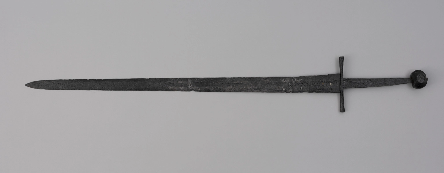miecz typu XIVa J2 (Oakeschott) - ujęcie z tyłu; Miecz długi półtoraręczny. Głownia długości 95 cm i szer. u nasady 5,6 cm, prosta dwusieczna zbieżna ku sztychowi, łączona wtórnie w dwóch miejscach. Zbrocze długie, sięga 3/4 długości. Pierwotnie po obu stronach głowni znajdował się napis ryty, wypełniony żółtym metalem w postaci krzyżyka oraz trzech liter, z których pierwsza jest nieczytelna, ale przypomina L lub T; druga, środkowa to D; trzecia litera to połączenie I i H. Trzpień rękojeści zwężający się ku końcowi. Jelec krzyżowy o długości 16,7 cm, z rozszerzającymi się ramionami. Głowica kolista, fazowana, dwubocznie spłaszczona o wys. 4,3 cm i szer. 5,6 cm. Powierzchnia z wżerami pokorozyjnymi. Typ wg Oakeschotta: XIVa J2 (ewentualnie XIIIa).