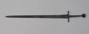 miecz typu XIVa J2 (Oakeschott) - ujęcie z tyłu; Miecz długi półtoraręczny. Głownia długości 95 cm i szer. u nasady 5,6 cm, prosta dwusieczna zbieżna ku sztychowi, łączona wtórnie w dwóch miejscach. Zbrocze długie, sięga 3/4 długości. Pierwotnie po obu stronach głowni znajdował się napis ryty, wypełniony żółtym metalem w postaci krzyżyka oraz trzech liter, z których pierwsza jest nieczytelna, ale przypomina L lub T; druga, środkowa to D; trzecia litera to połączenie I i H. Trzpień rękojeści zwężający się ku końcowi. Jelec krzyżowy o długości 16,7 cm, z rozszerzającymi się ramionami. Głowica kolista, fazowana, dwubocznie spłaszczona o wys. 4,3 cm i szer. 5,6 cm. Powierzchnia z wżerami pokorozyjnymi. Typ wg Oakeschotta: XIVa J2 (ewentualnie XIIIa).