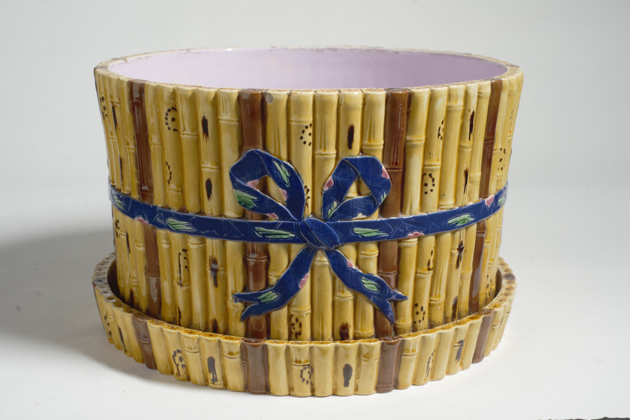 duże naczynie cylindryczne, o grubych ściankach, z podstawką. Zewnętrzna powierzchnia ścianek modelowana jest na kształt wiązek łodyg bambusowych przewiązanych wstążką z kokardą. Łodygi malowane są naturalistycznie, w kolorze żółtym i brązowym. Donica i podstawka pokryte są wewnątrz polewą w kolorze liliowym. 