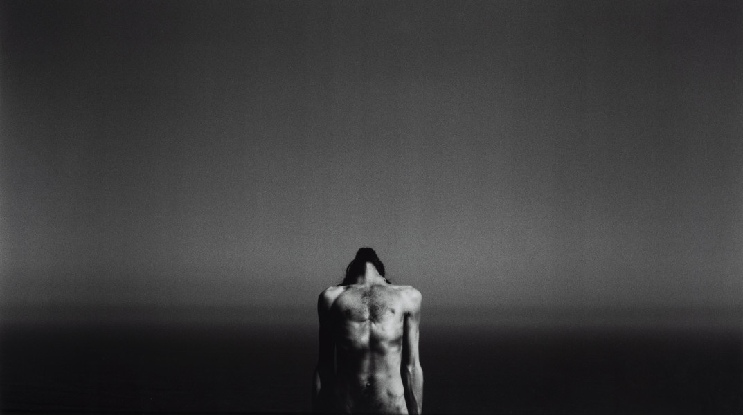 Z cyklu Rehearsals for Adulthood (The Dreamer, after Man Ray) - ujęcie z przodu; Jedna z dwóch czarno-białych fotografii oprawiona w białą drewnianą ramę. Pozioma praca przedstawia sylwetkę nagiego mężczyzny, od bioder w górę, wygiętego w tył, usytuowanego przodem do widza na tle spokojnego morza i linii horyzontu rozciągającej się mniej więcej na wysokości klatki piersiowej postaci i jednej trzeciej fotografii od dołu. Gładka tafla morza prawie zlewa się z horyzontem nieba. Na środku pracy, przy dolnej krawędzi jest postać z twarzą odchyloną ku górze.