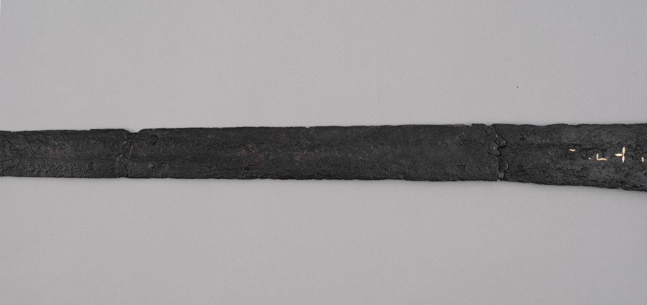 miecz typu XIVa J2 (Oakeschott) - ujęcie 3/4; Miecz długi półtoraręczny. Zbrocze długie, sięga 3/4 długości. Pierwotnie po obu stronach głowni znajdował się napis ryty, wypełniony żółtym metalem w postaci krzyżyka oraz trzech liter, z których pierwsza jest nieczytelna, ale przypomina L lub T; druga, środkowa to D; trzecia litera to połączenie I i H. Trzpień rękojeści zwężający się ku końcowi. Powierzchnia z wżerami pokorozyjnymi. Typ wg Oakeschotta: XIVa J2 (ewentualnie XIIIa).