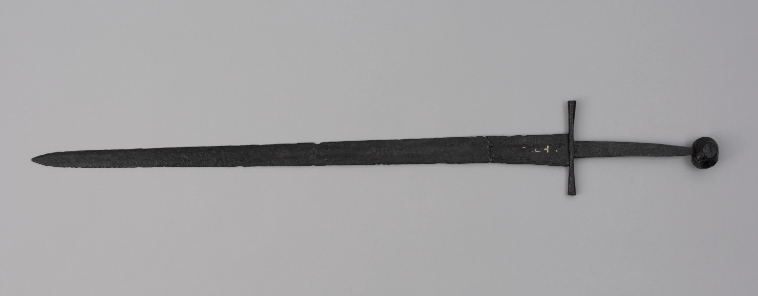 miecz typu XIVa J2 (Oakeschott) - ujęcie z przodu; Miecz długi półtoraręczny. Głownia długości 95 cm i szer. u nasady 5,6 cm, prosta dwusieczna zbieżna ku sztychowi, łączona wtórnie w dwóch miejscach. Zbrocze długie, sięga 3/4 długości. Pierwotnie po obu stronach głowni znajdował się napis ryty, wypełniony żółtym metalem w postaci krzyżyka oraz trzech liter, z których pierwsza jest nieczytelna, ale przypomina L lub T; druga, środkowa to D; trzecia litera to połączenie I i H. Trzpień rękojeści zwężający się ku końcowi. Jelec krzyżowy o długości 16,7 cm, z rozszerzającymi się ramionami. Głowica kolista, fazowana, dwubocznie spłaszczona o wys. 4,3 cm i szer. 5,6 cm. Powierzchnia z wżerami pokorozyjnymi. Typ wg Oakeschotta: XIVa J2 (ewentualnie XIIIa).