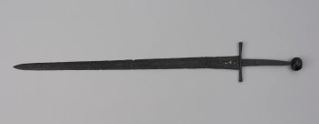 miecz typu XIVa J2 (Oakeschott) - ujęcie z przodu; Miecz długi półtoraręczny. Głownia długości 95 cm i szer. u nasady 5,6 cm, prosta dwusieczna zbieżna ku sztychowi, łączona wtórnie w dwóch miejscach. Zbrocze długie, sięga 3/4 długości. Pierwotnie po obu stronach głowni znajdował się napis ryty, wypełniony żółtym metalem w postaci krzyżyka oraz trzech liter, z których pierwsza jest nieczytelna, ale przypomina L lub T; druga, środkowa to D; trzecia litera to połączenie I i H. Trzpień rękojeści zwężający się ku końcowi. Jelec krzyżowy o długości 16,7 cm, z rozszerzającymi się ramionami. Głowica kolista, fazowana, dwubocznie spłaszczona o wys. 4,3 cm i szer. 5,6 cm. Powierzchnia z wżerami pokorozyjnymi. Typ wg Oakeschotta: XIVa J2 (ewentualnie XIIIa).