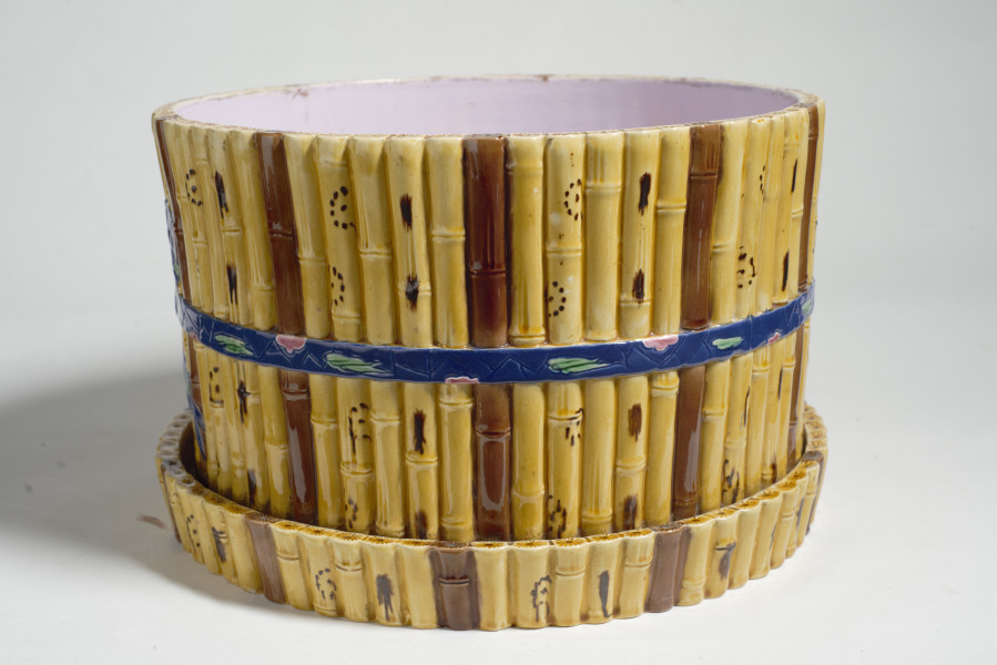 duże naczynie cylindryczne, o grubych ściankach, z podstawką. Zewnętrzna powierzchnia ścianek modelowana jest na kształt wiązek łodyg bambusowych przewiązanych wstążką z kokardą. Łodygi malowane są naturalistycznie, w kolorze żółtym i brązowym. Donica i podstawka pokryte są wewnątrz polewą w kolorze liliowym. 
