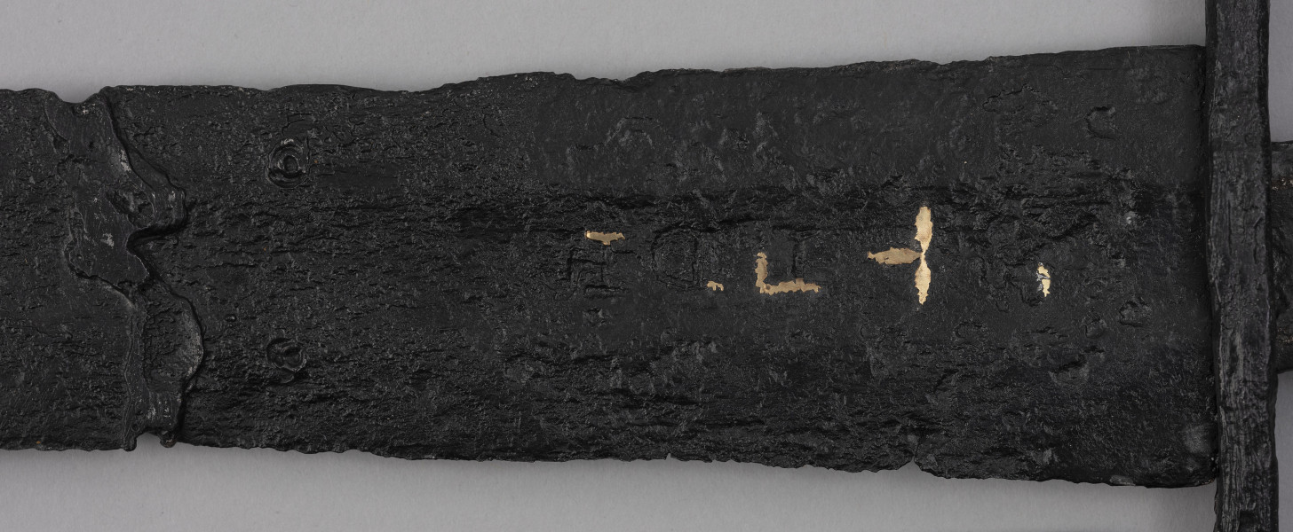 miecz typu XIVa J2 (Oakeschott) - detal; Miecz długi półtoraręczny. Fragment zbrocza. Pierwotnie po obu stronach głowni znajdował się napis ryty, wypełniony żółtym metalem w postaci krzyżyka oraz trzech liter, z których pierwsza jest nieczytelna, ale przypomina L lub T; druga, środkowa to D; trzecia litera to połączenie I i H.