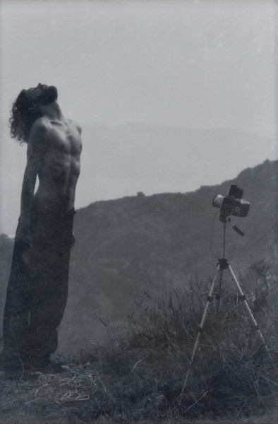 Z cyklu Rehearsals for Adulthood (The Dreamer, after Man Ray) - ujęcie z tyłu; Jedna z dwóch czarno-białych fotografii. Fotografia pionowa przedstawia całą sylwetkę mężczyzny z nagim torsem, w ciemnych spodniach, ustawioną pod kątem do widza, z lewej strony pracy. Na wprost mężczyzny znajduje się aparat fotograficzny na statywie/trójnogu ustawionego w trawie, na łące z widokiem gór w tle. Ręce postaci są wyprostowane wzdłuż tułowia, głowa mocno odchylona w tył, twarz zwrócona ku niebu. Można się domyślić, ze na zdjęciu b widzimy samego artystę, który robi sobie zdjęcie; za nim musi znajdować się morze, które jest tłem i jednocześnie ważnym. dominującym elementem fotografii pierwszej.