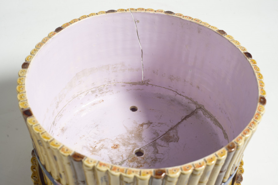 cache-pôt to duże naczynie cylindryczne, o grubych ściankach, z podstawką. Zewnętrzna powierzchnia ścianek modelowana jest na kształt wiązek łodyg bambusowych przewiązanych wstążką z kokardą. Łodygi malowane są naturalistycznie, w kolorze żółtym i brązowym. Donica i podstawka pokryte są wewnątrz polewą w kolorze liliowym. 