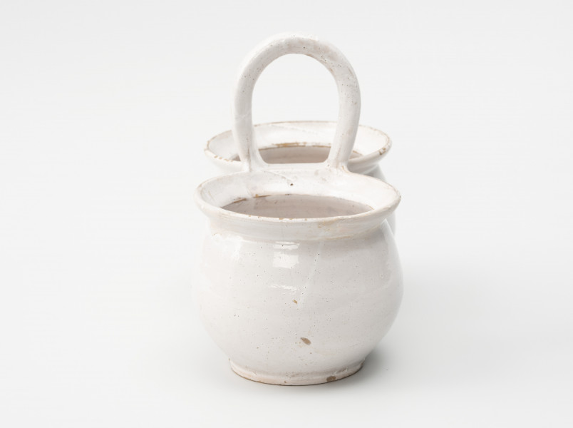 dwojaki - Ujęcie z boku; Szkliwione na biało naczynie składa się z dwóch jednakowych pojemników w formie garnków o pękatych brzuścach, przechodzących w bardzo krótką i nieznacznie tylko przewężoną szyjkę, a następnie w wylew z wyraźnie zaznaczonymi szerokimi kołnierzami. Naczynia łączą się ze sobą górną krawędzią wylewów, do których doklejono kabłąkowe, poprzecznie osadzone ucho.