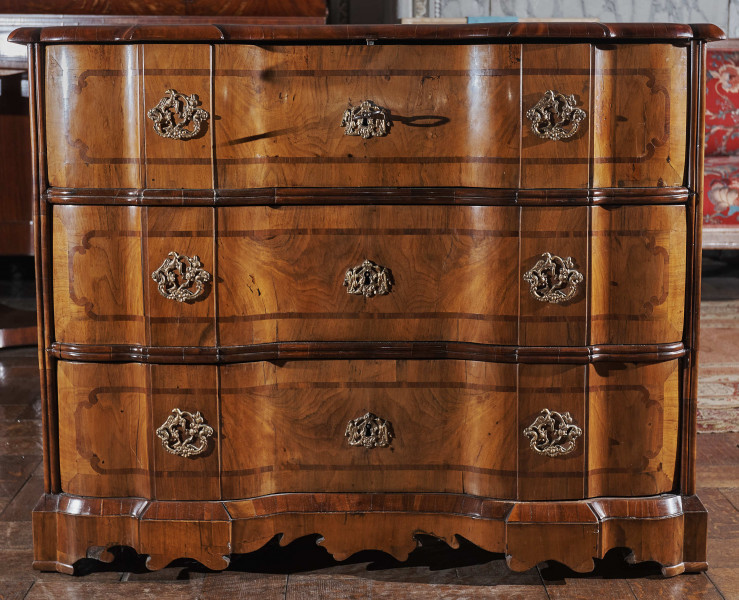 Posiada proste boki i falujący front z trzema szufladami. Przednie nogi konsolowe przechodzą w dekoracyjnie wycięty fartuch, który przesłania prześwit pod meblem. Fornir intarsjowany wykonano z różnych odmian orzecha i mahoniu. Lica szuflad, boki i blat zdobione intarsją w postaci zgeometryzowanej wstęgi. Blat obwiedziony dekoracyjnym ćwierćwałkiem z uskokami. Pomiędzy szufladami zastosowano poziome, rozdzielające półwałki. Szuflady posiadają brązowe okucia ozdobione rocaillami. Każda z nich posiada dwa uchwyty po bokach oraz ozdobne mniejsze okucie na środku przysłaniające dziurkę od klucza.