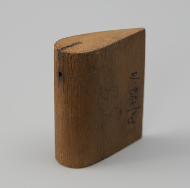 tłok pieczętny - Ujęcie z tyłu; Bierka płaska wykonana z jednego kawałka drewna. W przekroju ma kształt łezkowaty, płaszczyznę profilowaną. Góra i dół ścięte są pod kątem prostym, przód i tył są zaoblone. Na grubszym końcu, z przodu w części zaoblonej otwór, a drewno w tych miejscach jest nieco przyciemnione.