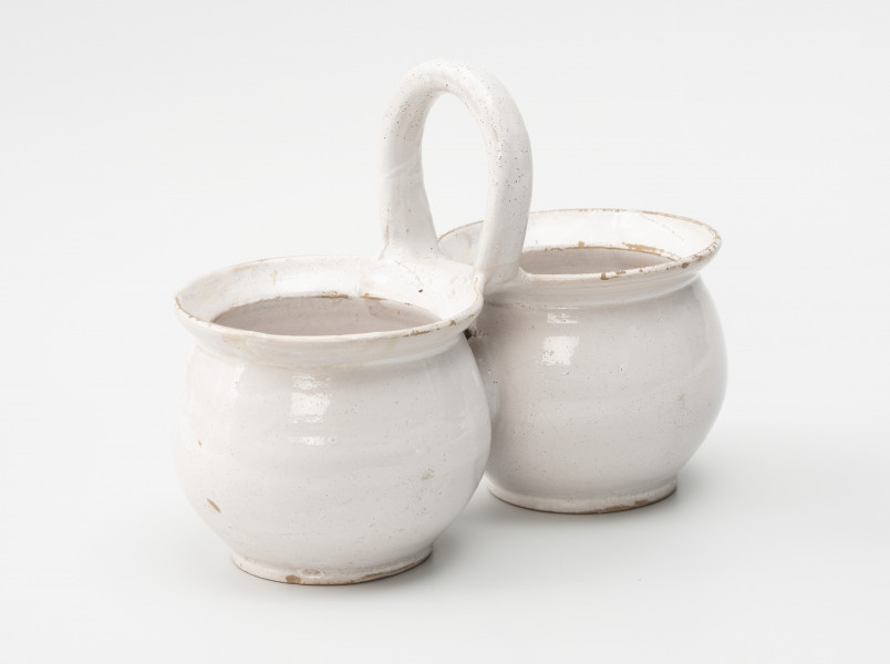 dwojaki - Ujęcie ze skosu; Szkliwione na biało naczynie składa się z dwóch jednakowych pojemników w formie garnków o pękatych brzuścach, przechodzących w bardzo krótką i nieznacznie tylko przewężoną szyjkę, a następnie w wylew z wyraźnie zaznaczonymi szerokimi kołnierzami. Naczynia łączą się ze sobą górną krawędzią wylewów, do których doklejono kabłąkowe, poprzecznie osadzone ucho. Brzuśce połączono za pomocą wałka gliny.