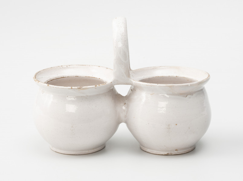 dwojaki - Ujęcie z przodu; Szkliwione na biało naczynie składa się z dwóch jednakowych pojemników w formie garnków o pękatych brzuścach, przechodzących w bardzo krótką i nieznacznie tylko przewężoną szyjkę, a następnie w wylew z wyraźnie zaznaczonymi szerokimi kołnierzami. Naczynia łączą się ze sobą górną krawędzią wylewów, do których doklejono kabłąkowe, poprzecznie osadzone ucho. Brzuśce połączono za pomocą wałka gliny.