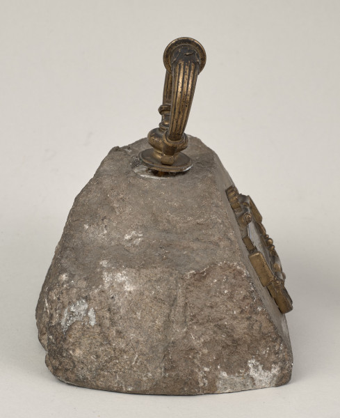 Przycisk do papieru w kształcie leżącego kamiennego nieforemnego prostopadłościanu o nieregularnych krawędziach na górze ma zamontowanym okrągłym mosiężnym uchwyt, z tego samego materiału wykonana jest podłużna ozdobna tabliczka ozdobiona motywem muszli mocowana do kamienia bez widocznych ćwieków z napisem „Stare Sioło 1872