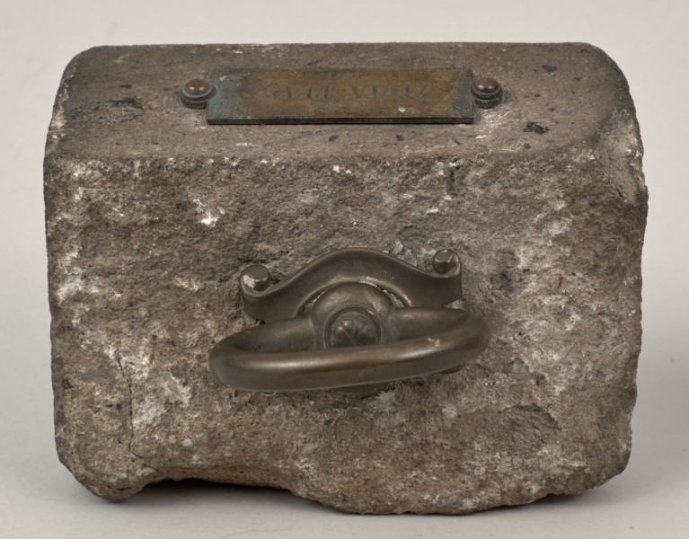 Przycisk do papieru w kształcie leżącego kamiennego prostopadłościanu o lekko zaokrąglonych krawędziach, na górze ma zamontowany okrągły mosiężny uchwyt na stylizowanym kartuszu, z  tego samego materiału wykonana jest podłużna tabliczka mocowana do kamienia dwoma ćwiekami z napisem „Kijów – Złota Brama 1871