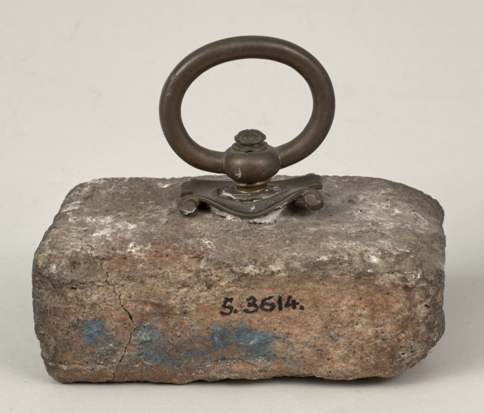 Przycisk do papieru w kształcie leżącego kamiennego prostopadłościanu o lekko zaokrąglonych krawędziach, na górze ma zamontowany okrągły mosiężny uchwyt na stylizowanym kartuszu, z  tego samego materiału wykonana jest podłużna tabliczka mocowana do kamienia dwoma ćwiekami z napisem „Kijów – Złota Brama 1871