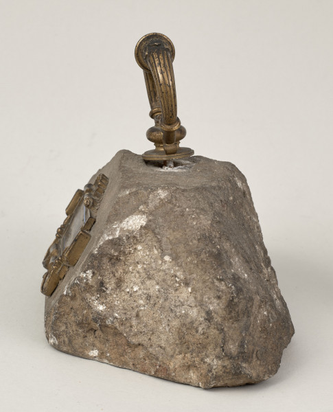 Przycisk do papieru w kształcie leżącego kamiennego nieforemnego prostopadłościanu o nieregularnych krawędziach na górze ma zamontowanym okrągłym mosiężnym uchwyt, z tego samego materiału wykonana jest podłużna ozdobna tabliczka ozdobiona motywem muszli mocowana do kamienia bez widocznych ćwieków z napisem „Stare Sioło 1872