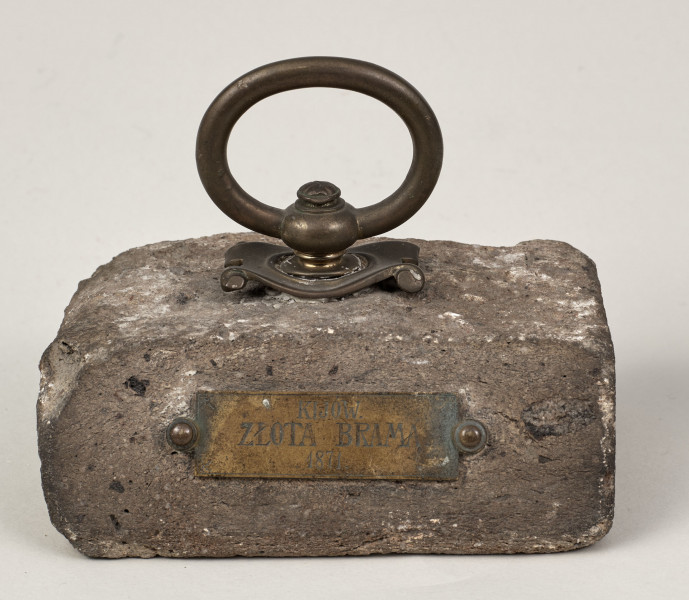 Przycisk do papieru w kształcie leżącego kamiennego prostopadłościanu o lekko zaokrąglonych krawędziach, na górze ma zamontowany okrągły mosiężny uchwyt na stylizowanym kartuszu, z  tego samego materiału wykonana jest podłużna tabliczka mocowana do kamienia dwoma ćwiekami z napisem „Kijów – Złota Brama 1871