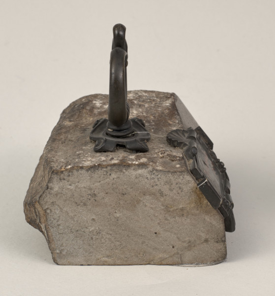 Przycisk do papieru w kształcie leżącego kamiennego nieforemnego prostopadłościanu o  szlifowanych bokach i ścianie z tabliczką, na górze ma zamontowany okrągły mosiężny uchwyt z tego samego materiału wykonana jest podłużna ozdobna tabliczka ozdobiona motywem muszli, mocowana do kamienia bez widocznych ćwieków, z napisem „Trembowla - 1870”