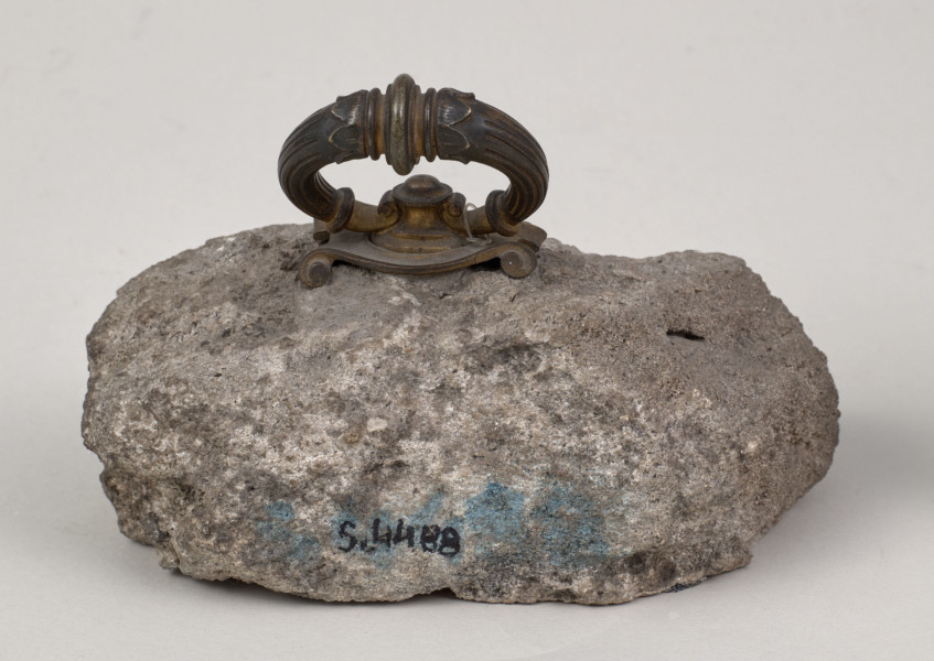 Przycisk w kształcie leżącego kamiennego nieforemnego prostopadłościanu o nieregularnych krawędziach, na górze ma zamontowany okrągły mosiężny z tego samego materiału wykonana jest podłużna ozdobna tabliczka ozdobiona motywem muszli  i mocowana do kamienia bez widocznych ćwieków z napisem „Zamek Krzemieniecki – 1871”. 