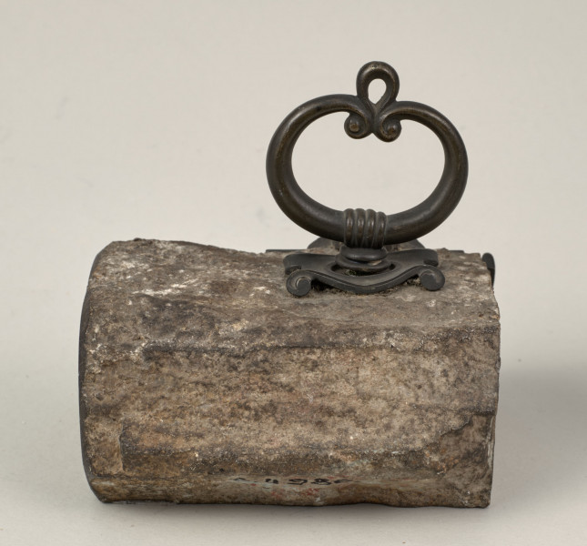 Przycisk do papieru w kształcie leżącego kamiennego nieforemnego prostopadłościanu o  szlifowanych bokach i ścianie z tabliczką, na górze ma zamontowany okrągły mosiężny uchwyt z tego samego materiału wykonana jest podłużna ozdobna tabliczka ozdobiona motywem muszli, mocowana do kamienia bez widocznych ćwieków, z napisem „Trembowla - 1870”