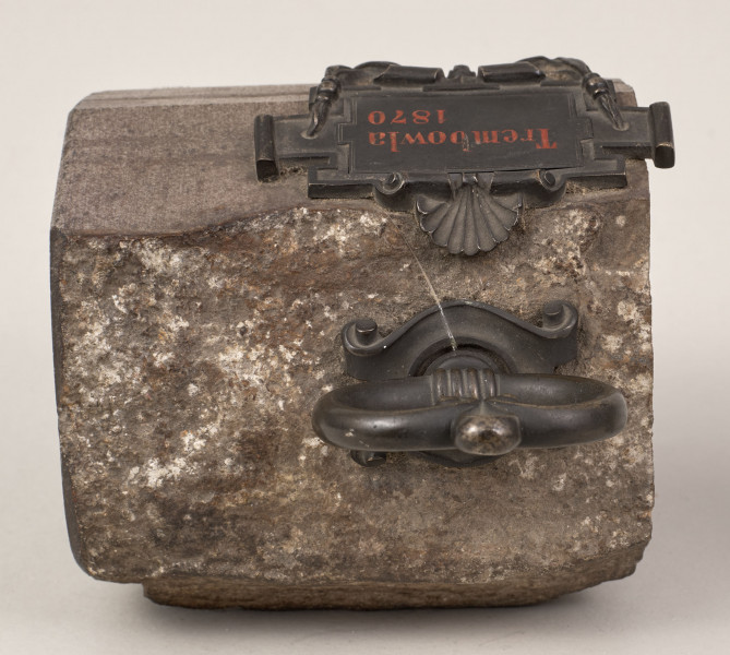 Przycisk do papieru w kształcie leżącego kamiennego nieforemnego prostopadłościanu o  szlifowanych bokach i ścianie z tabliczką, na górze ma zamontowany okrągły mosiężny uchwyt z tego samego materiału wykonana jest podłużna ozdobna tabliczka ozdobiona motywem muszli, mocowana do kamienia bez widocznych ćwieków, z napisem „Trembowla - 1870”