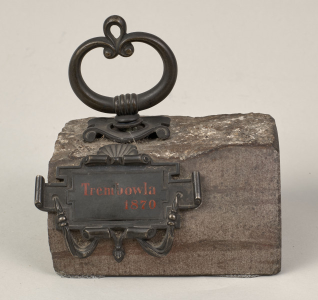 Przycisk do papieru w kształcie leżącego kamiennego nieforemnego prostopadłościanu o  szlifowanych bokach i ścianie z tabliczką, na górze ma zamontowany okrągły mosiężny uchwyt z tego samego materiału wykonana jest podłużna ozdobna tabliczka ozdobiona motywem muszli, mocowana do kamienia bez widocznych ćwieków, z napisem „Trembowla - 1870”