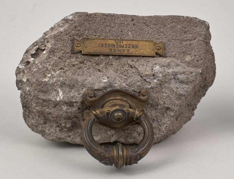 Przycisk w kształcie leżącego kamiennego nieforemnego prostopadłościanu o nieregularnych krawędziach, na górze ma zamontowany okrągły mosiężny z tego samego materiału wykonana jest podłużna ozdobna tabliczka ozdobiona motywem muszli  i mocowana do kamienia bez widocznych ćwieków z napisem „Zamek Krzemieniecki – 1871”. 