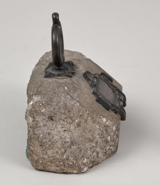 Przycisk do papieru w kształcie leżącego kamiennego nieforemnego prostopadłościanu o  szlifowanych jednym boku, drugi nieforemnie zaokrąglony i ścianie z tabliczką lekko wygładzona na górze ma zamontowany okrągły mosiężny uchwyt z tego samego materiału wykonana jest podłużna ozdobna tabliczka ozdobiona motywem muszli mocowana do kamienia bez widocznych ćwieków mocujących z napisem „Sobieskich Złoczów - 1870”. 