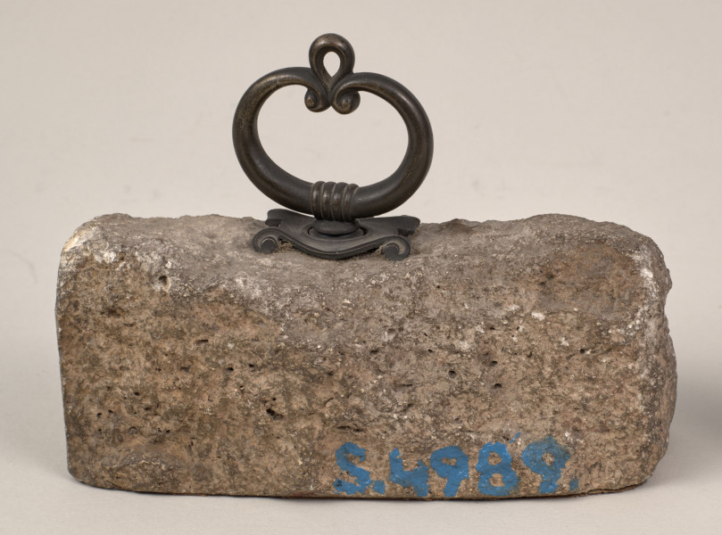 Przycisk do papieru w kształcie leżącego kamiennego nieforemnego prostopadłościanu o  szlifowanych jednym boku, drugi nieforemnie zaokrąglony i ścianie z tabliczką lekko wygładzona na górze ma zamontowany okrągły mosiężny uchwyt z tego samego materiału wykonana jest podłużna ozdobna tabliczka ozdobiona motywem muszli mocowana do kamienia bez widocznych ćwieków mocujących z napisem „Sobieskich Złoczów - 1870”. 