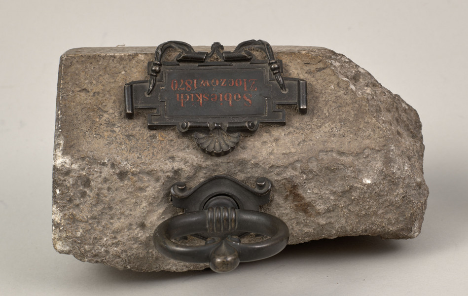 Przycisk do papieru w kształcie leżącego kamiennego nieforemnego prostopadłościanu o  szlifowanych jednym boku, drugi nieforemnie zaokrąglony i ścianie z tabliczką lekko wygładzona na górze ma zamontowany okrągły mosiężny uchwyt z tego samego materiału wykonana jest podłużna ozdobna tabliczka ozdobiona motywem muszli mocowana do kamienia bez widocznych ćwieków mocujących z napisem „Sobieskich Złoczów - 1870”. 