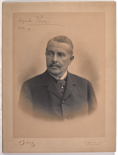 Fotografia przedstawia mężczyznę w średnim wieku ukazanym w popiersiu, en face z głową skierowaną w lewo i wzrokiem skierowanym przed siebie