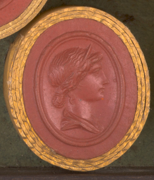 czerwona gemma, owalna pionowa, w grubym złotym obramowaniu. Prawy profil młodej kobiety o spiętych na karku włosach i diademie na głowie.