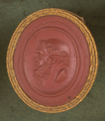 czerwona owalna gemma w grubym złotym obramowaniu; lewy profil głowy starszego, łysego mężczyzny z gęstą kręconą brodą i wąsami, na głowie ma wieniec z winorośli 