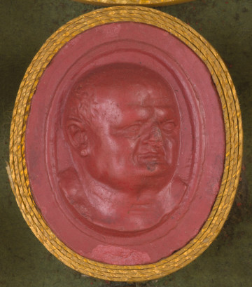 czerwona owalna gemma w grubym złotym obramowaniu; głowa starszego mężczyzny obrócona lekko w lewo. Mężczyzna ma okrągłą głowę, szeroką twarz i wystający podbródek, łysieje - krótkie włosy widoczne jedynie na skroniach