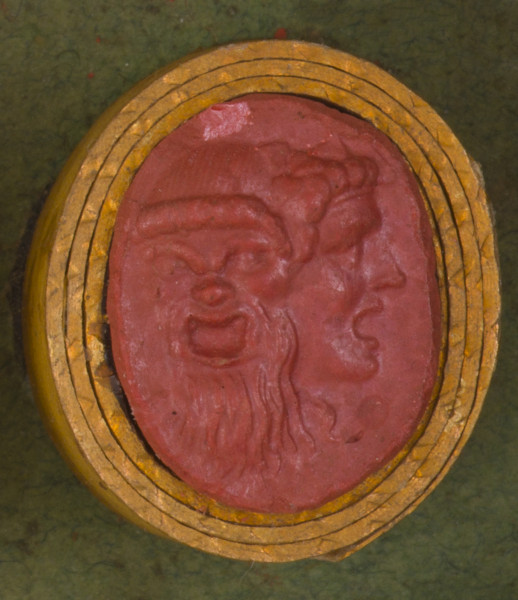 czerwona owalna gemma w grubym złotym obramowaniu; dwie maski groteskowe połączone razem, maska po lewej przedstawia brodatego starca, maska po prawej młodzieńca