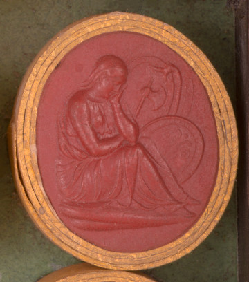 czerwona gemma, owalna pionowa, w grubym złotym obramowaniu. Prawy profil siedzącej na ziemi kobiety, w długiej szacie i chuście na głowie. Nogi zgiete w kolanach, głowa wsparta na lewej dłoni. W tle tarcza i topór.