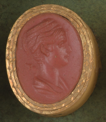 czerwona owalna gemma w grubym złotym obramowaniu; młoda kobieta widziana z prawego profilu, ma długie włosy zaplecione w warkocz, który został owinięty wok~ł jej głowy, a z tyłu są zpięte w kok; z tyłu, koło lewego ramienia, widoczna rękojeść miecza, poniżej fragment szaty