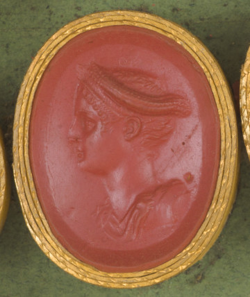 czerwona owalna gemma w grubym złotym obramowaniu; popiersie młodej kobiety widoczne z lewego profilu, kobieta ma dekoracyjne nakrycie głowy, poniżej wida fragment szaty