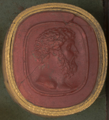 czerwona owalna gemma w grubym złotym obramowaniu; prawy profil głowy mężczyzny w średnim wieku, ma on kręcone krótkie włosy, brodę i wąsy, jest dobrze zbudowany, o czym świadczy gruba szyja; widoczne wąskie ukośne pęknięcie biegnące przez całą gemmę