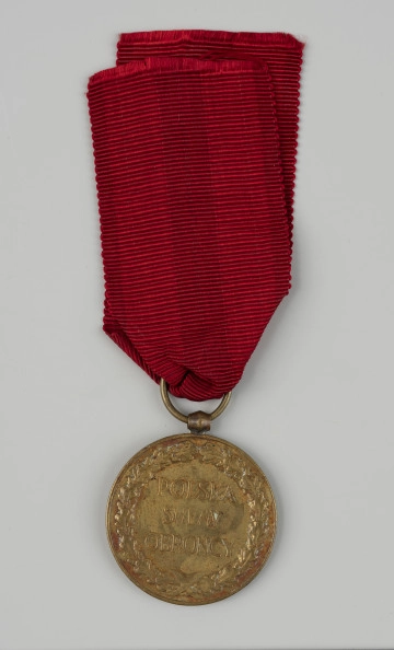 MNS/CDP-p/416/2 Medal Pamiątkowy Za Wojnę 1918-1921 - rewers; Okrągły medal z oczkiem do zawieszania na górze. W polu rewersu znajduje się dewiza w trzech wierszach: POLSKA SWEMU OBROŃCY, otoczona wieńcem z liści dębowych. Medal zawieszony na zastępczej wstążce w kolorze czerwonym.